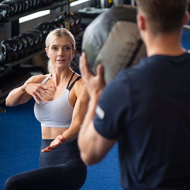 A personal trainer holds a weighted pad while a female client in athletic wear performs a rotational core exercise in a gym with dumbbell racks in the background.
