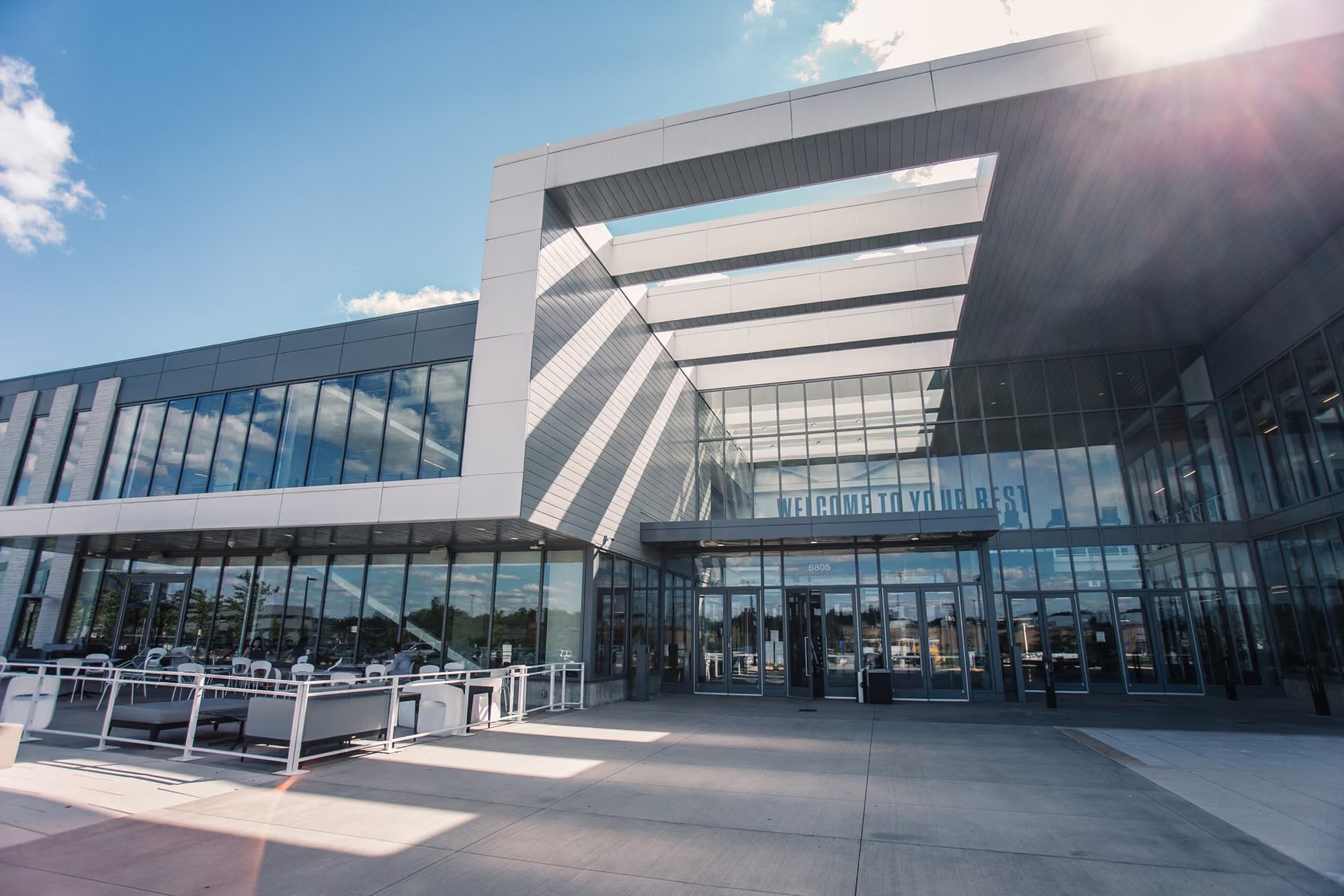Modern glass and metal building exterior with an angled entrance canopy, outdoor seating area, and 'Welcome to Your Rest' signage above the doors.