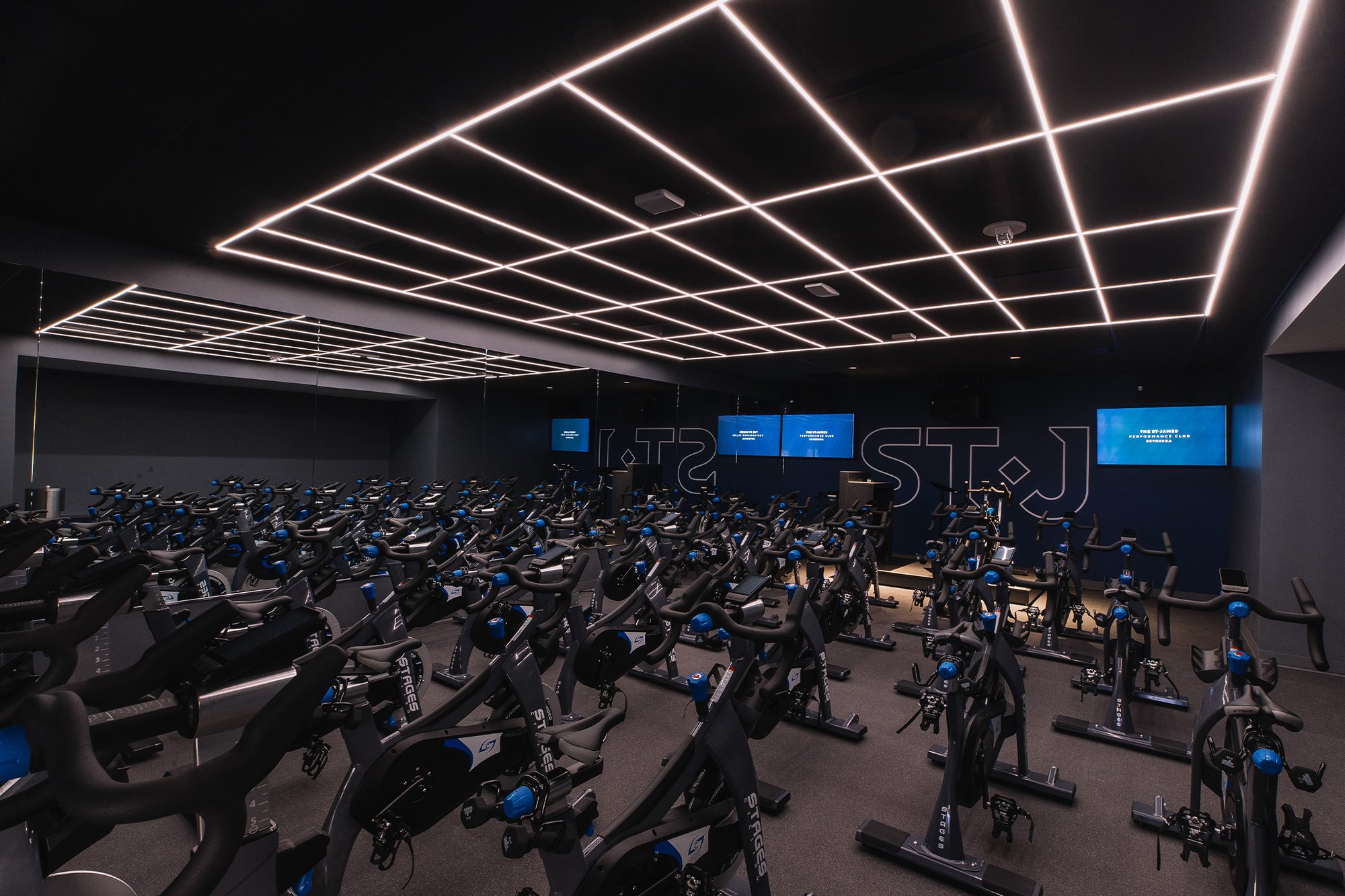 A spacious indoor cycling studio at The St. James Bethesda, lined with rows of stationary bikes under a striking grid-patterned LED ceiling and wall-mounted screens.