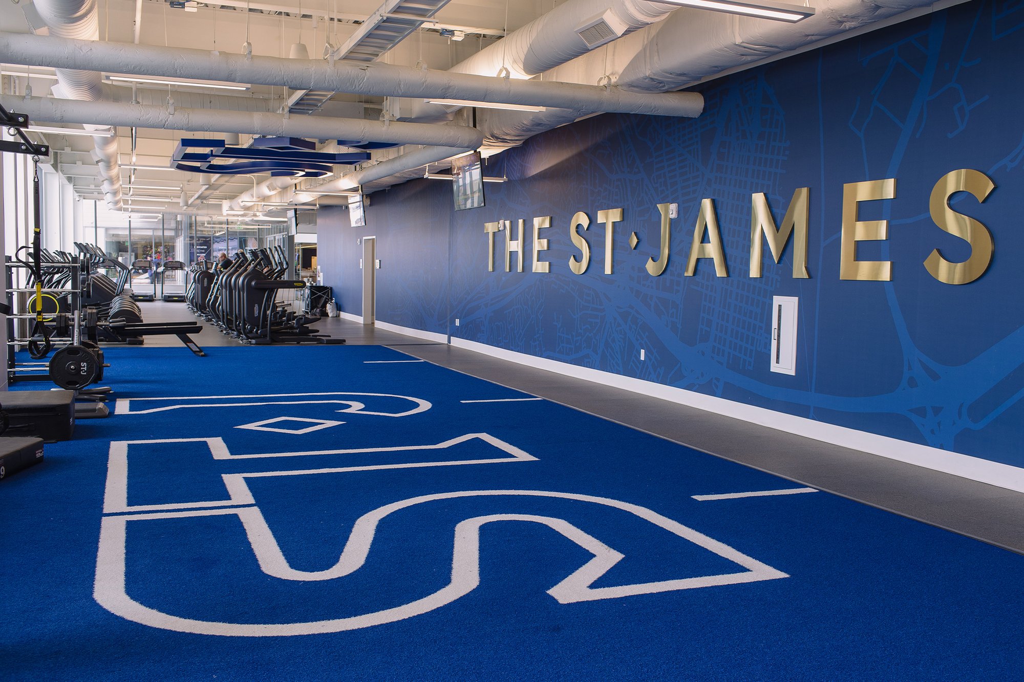 A bright open training floor at The St. James Northern Virginia, with the club's logo on blue turf flooring, treadmills, and gold signage reading 'The St. James' on the wall.